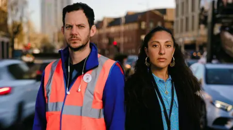 Tom Arran/Middle Child Two people stand side by side, a man is on the left, dressed in a high-vis work jacket and headphones and on the right, there is a solemn looking woman, wearing a smart shirt, dark coat and identification badge.