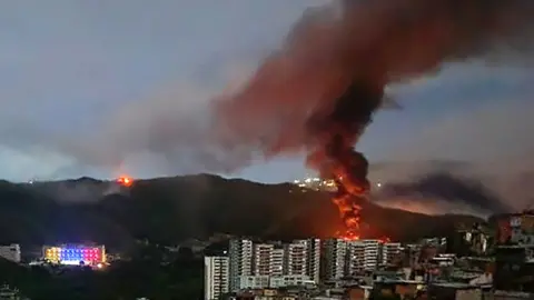 Smoke billows over Caracas, Venezuela with buildings in the foreground and a mountain in the background.