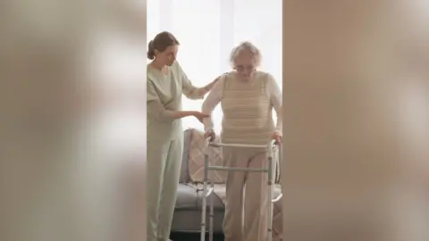 A woman in scrubs helps an older woman walk with a frame