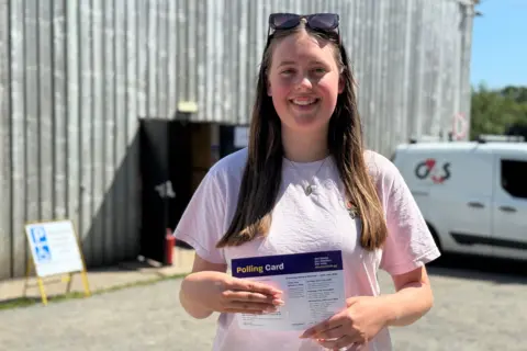 A young women with long brown hair smiling at the camera. She is wearing a pale pink top and has sunglasses resting on her head. She is hold her polling card up to the camera.