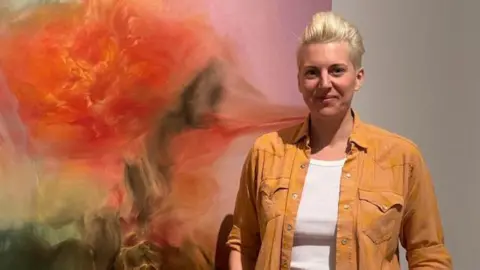 BBC/Emma Richardson A smiling Emma Richardson is wearing a camel coloured overshirt with a white t-shirt underneath. She's standing in front of her own oil painting which merges red, orange, green paint colours in a striking, largely blurred pattern. 