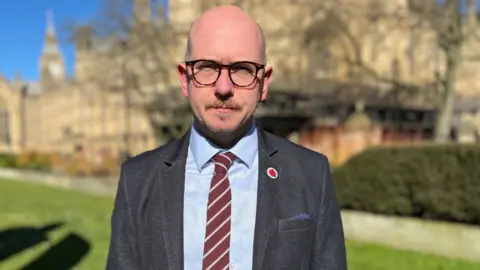 A bald man with glasses outside the Westminster Parliament on a sunny day. He is looking at the camera and is wearing a grey suit, blue shirt and maroon and grey striped tie. 