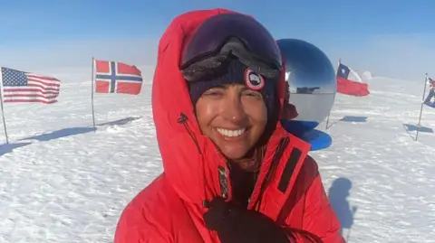 Harpreet Chandi wears a red coat and smiles to the camera. Snow is visible in the background with flags including those of the USA and Norway in the background.