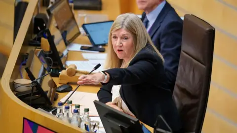 PA Media Alison Johnstone, who has long fair hair, points with her left hand outstretched while sitting in a leather chair at a wooden podium. 