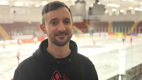 James Assinder, he is in the stands of an ice hockey arena and the rink is visible behind him. He has short brown hair and a light beard and is wearing a black hoodie with the logo of a dragon on it which is coloured orange and yellow