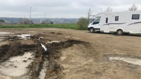 JUSTIN DEALEY/BBC A close up of the deep tracks in the mud left by a refuse lorry that had to be pulled out of the mud. A white motorhome, with black stripes on its side, is parked next to it.
