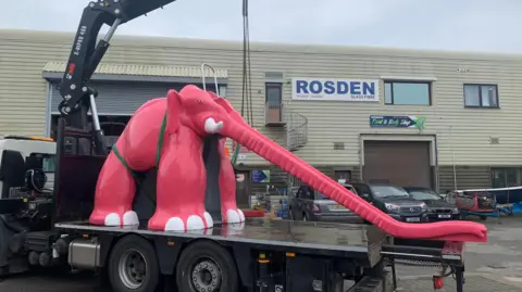 Chris Orkizewski A giant pink glass fibre elephant slide on a flatbed lorry against an industrial estate backdrop.