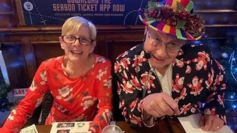 Geoff King A smiling Val and Geoff King are pictured during a pub quiz. She is wearing a red flowery Christmas dress. He is wearing a colourful hat and a suit jacket with a Father Christmas print. 