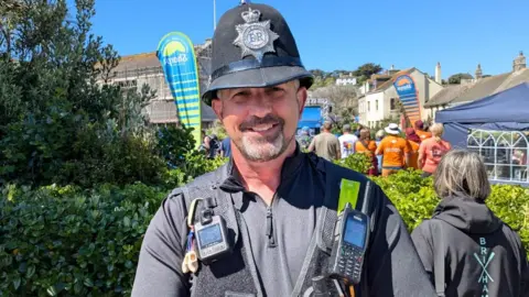 A UK police officer with a beard and traditional hat in black uniform with a number of devices attached.