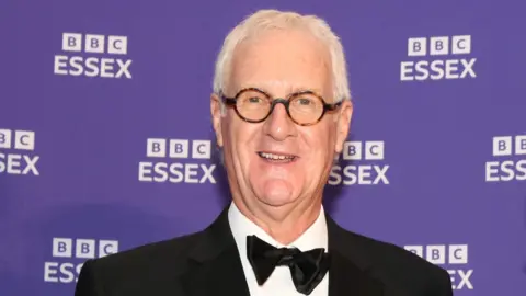 Dave Monk stands in front of a purple backdrop with several white BBC Essex logos. Dave is wearing a tuxedo jacket, white shirt and black bow tie. He has grey hair, is smiling at the camera and wears round spectacles.