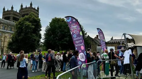Durham Fringe People queueing outside a Durham Fringe tent on the city's Palace Green outside Durham Cathedral in July 2023. there are purple Durham Fringe banners.
