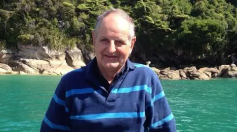 Robert was a man in his late-middle years with thinning grey hair. He is wearing a deep and light blue striped overshirt and is in front of brilliant green water, with a rocky shore and some woods in the background.