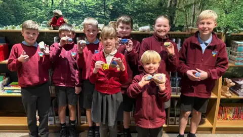 Eight children in a school room smiling at the camera. They are wearing burgundy jumpers and sweatshirts and are holding painted rocks in their hands. They are standing in front of wooden book shelves and wallpaper resembling a green forest.