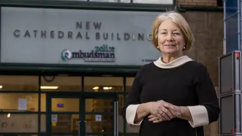 PA Media Marie Anderson standing outside the police ombudsman office in Belfast. She wears a black dress with a white collar and cuffs. She has short, blonde hair.