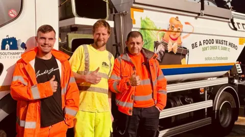 Three men in hi-viz clothing pose for a picture in front of a large food waste collection vehicle. The men are all raising their right thumb for the photo. 
