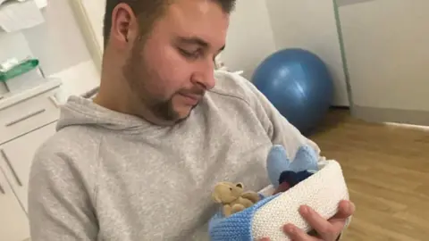 A man in a grey hoodie cradles a tiny stillborn baby in a knitted blue and white basket, along with a small beige teddy and a blue felt heart.