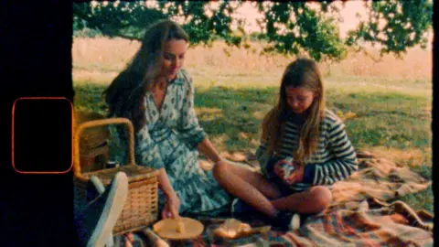 Will Warr/Kensington Palace Catherine and Princess Charlotte sit together on a picnic blanket alongside food. Charlotte looks to be shuffling a deck of cards in the scene which is treated to appear like it is being played on old film