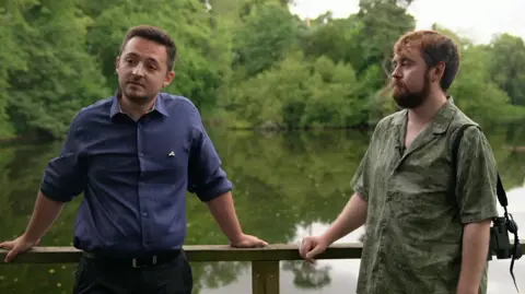 Ryan and Adam stand together leaning on a wooden railing by a lake. Ryan has a blue shirt on and dark trousers. He has short brown hair. Adam has a green shirt on with a camera over his shoulder. He has red hair and a beard.