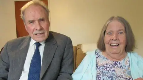 Francis Hillier and his wife Janice Hillier pictured on their 60th wedding anniversary in 2025. They are sitting down on a brown couch.