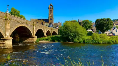 BBC Weather Watchers - Alan Historic stone arch bridge crossing a shallow river with flowing water, leading toward a town with buildings and a tall church tower under a bright blue sky, surrounded by green trees.