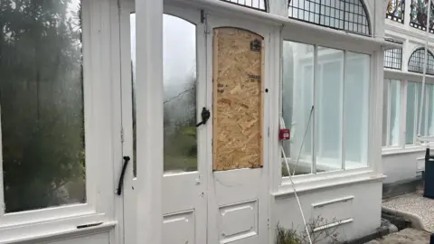 The doors inside a light conservatory. The wall is lined with windows, and one set of double doors. One of the doors has it's window blocked with a wooden board. Below the windows, the wall is white and there is a white tiled floor. 