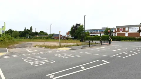 Donna Clifford The site of the new car park overgrown with weeds located on the corner of a junction with traffic lights with the red brick, two-storey building of the school in the background