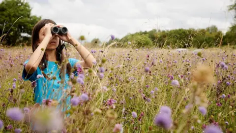 Oliver Edwards Photography Mya-Rose stands in a field with purple flowers. She is holding a pair of binoculars up to her eyes as she spots birds in the distance. She is wearing a blue top and has long brown hair.