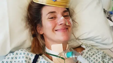 SARRA WILSON Sarra Wilson in hospital in 2018. This was the first photograph her daughter saw of her after her accident, so she smiled for the camera and is wearing a paper crown from a fast food takeaway. She has a trachea on her neck and has a tubed taped to her nose, she is laying in bed wearing a hospital gown.