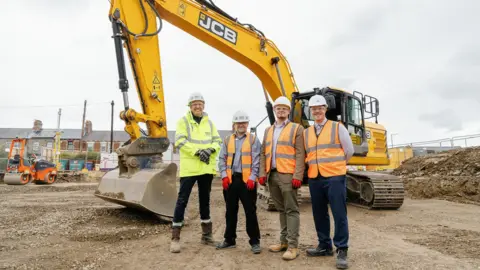 Durham County Council Four men in high vis jackets and hard hats stand in front of a large yellow JCB digger on a construction site. The land has been cleared and is muddy, with a row of houses beyond.