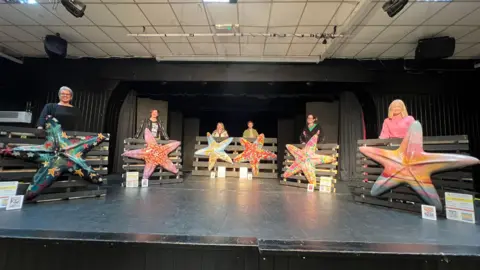 A black stage with six people each standing behind large starfish sculptures. They are all individually designed and very colourful.