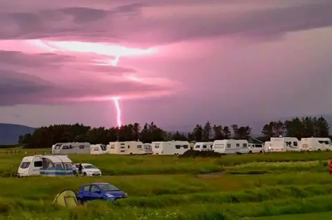 Gareth Hall A pink sky over Dornoch Caravan and Camping Park with a bright lightning bolt. There are caravans in the grassy field and clouds in the sky. 