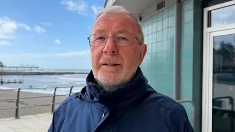 A photo of Gareth Davies - bearded with glasses and a blue coat - standing on Aberystwyth promenade with the pier and blue sky in the background