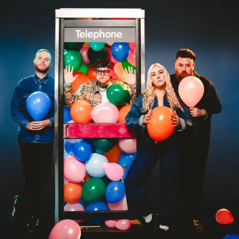 Lottery Winners Thom Rylance is standing in a phone box filled with balloons. The other band members - Joe Singleton, Katie Lloyd and Robert Lally - are standing outside the box holding different coloured balloons.