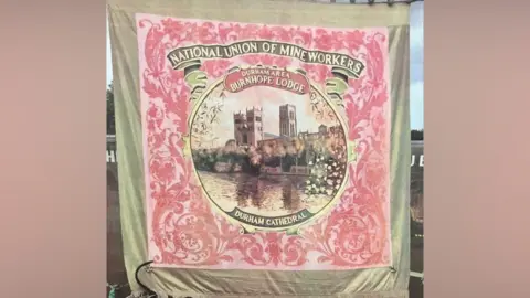 Supplied Burnhope Miners' Banner is gold and pink. It shows Durham Cathedral and reads NATIONAL UNION OF MINEWORKERS DURHAM AREA BURNHOPE lODGE DURHAM CATHEDRAL