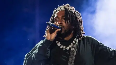 Rapper Kendrick Lamar, wearing black clothes, a necklace, and a bag over his shoulder, holds a microphone as he performs onstage during day three of Rolling Loud Miami 2022 at Hard Rock Stadium on July 24, 2022 in Miami Gardens, Florida