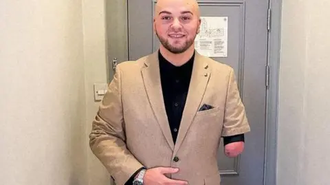 Ryan Rix Ryan Rix who is bald with a beard and one forearm, wearing a light brown suit, standing in front of a grey door