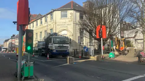 A large lorry parked on a road next to the run down looking white citadel building with a sign that reads The Salvation Army Citadel above the entrance. The temporary lights have been set up next to the building. Two men are working by a nearby yellow digger and there is a blue sky above.