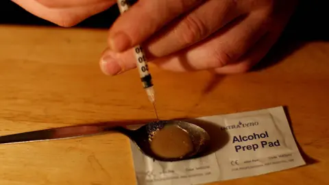 A hand holding a syringe, drawing a brown liquid substance into the syringe from a metal spoon.