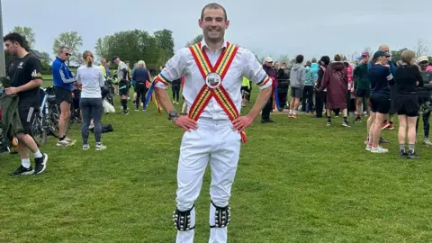 A man with short brown hair in a field dressed in Morris dancing garb.