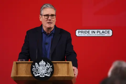 A man with grey hair and glasses, wearing a blue shirt, standing infront of a bright red background