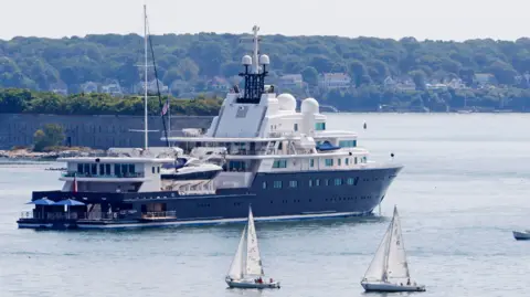 Getty Images Le Grand Bleu, sebuah kapal pesiar besar dengan lunas biru tua dan geladak atas putih, dengan payung yang terlihat di platform renang besar di bagian belakang, berlabuh di dekat Portland di Maine, AS, dengan kapal layar kecil lewat.