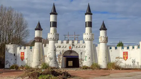 Alamy The entrance to the abandoned Camelot theme park. The image shows a mock white castle frontage with a barbican and turrets. There are coats of arms on the walls and overgrowth in the foreground.