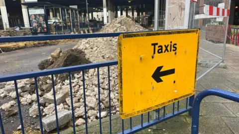 A sign attached onto fencing at Great Yarmouth Market Gates bus station on Temple Road, directs passengers to the nearest taxi ranks: Theatre Plain (outside Costa Coffee) and Market Gates Road. Behind the fencing is a pile of rubble on the roadway and the Market Gates shopping centre.