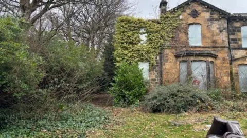 A building with boarded-up windows and overgrown bushes. There is a grassed area with an overturned chair.