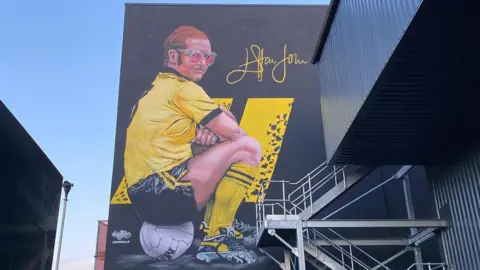 MurWalls A mural of Sir Elton John on the side of a football stadium. He wears a yellow football shirt and black shorts while sitting on a football. He has pink glasses and short ginger hair.