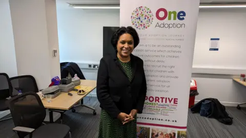 David Spereall/BBC Children's minister Janet Daby, a lady with dark hair, a black jacket and a green and black dress. She is stood partially in front of an advertising board for One Adoption, which has large writing on explaining what the agency is. A desk with various items is over her right shoulder.