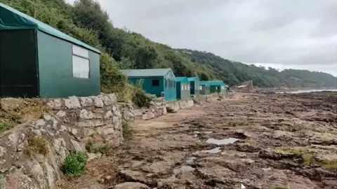 A row of sturdy green buildings on hard standing. They have pitched roofs and windows. There is a low wall in front of them and rocks leading out to the sea.