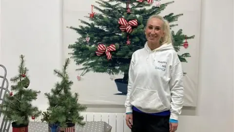 BBC/Caroline Wordsworth Michelle Clark, the founder of Happy Me Dance, standing in a white jumper and in front of a Christmas tree. She is smiling and looking directly at the camera. 