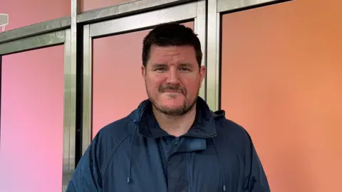 A brown haired male wearing a navy rain jack stares at the camera