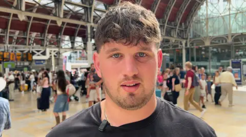 Man in black T-shirt with beard at busy station